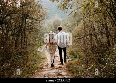 Vue arrière de jeune mariée et marié méconnaissable dans un style élégant ensemble de mariage marchant sur un chemin rocailleux étroit en montagne Forêt de Morro de Labella en Espagne Banque D'Images