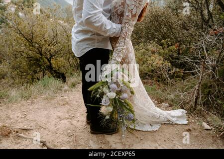 Vue latérale d'un petit couple romantique, jeune couple récemment marié et méconnu en tenues de mariage avec bouquet embrassant et embrassant tout en étant debout Dans le paysage pittoresque de montagne de Morro de Labella en Espagne Banque D'Images