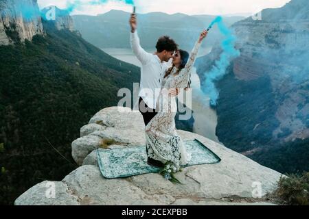 Vue latérale complète du jeune couple heureux et récemment marié dans des tenues élégantes tenant des bombes de fumée colorées et embrassant tout en Célébration du mariage sur le rocher de Morro de labella En Espagne Banque D'Images