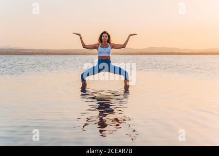 Pleine longueur de jeune femme mince en forme d'activewear debout à l'intérieur Déesse yoga asana dans l'eau calme du lac pendant le coucher du soleil d'été Banque D'Images