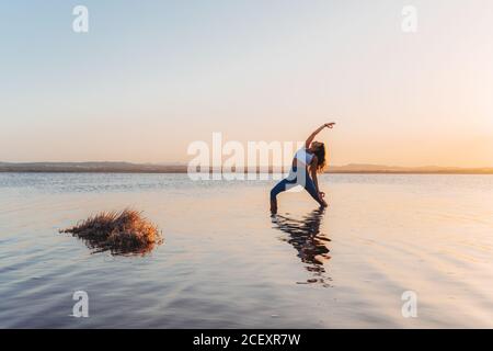 Pleine longueur de jeune femme mince en forme d'activewear debout à l'intérieur Posture du guerrier inversé dans l'eau calme du lac pendant le coucher du soleil d'été Banque D'Images