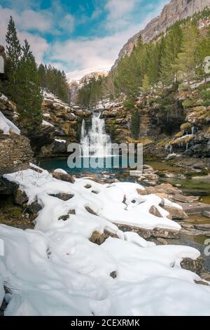 Paysage pittoresque de la rivière de montagne avec eau claire bleue en continu par la vallée entourée de montagnes rocheuses couvertes de neige et forêt verte en hiver ensoleillé avec ciel bleu nuageux Banque D'Images