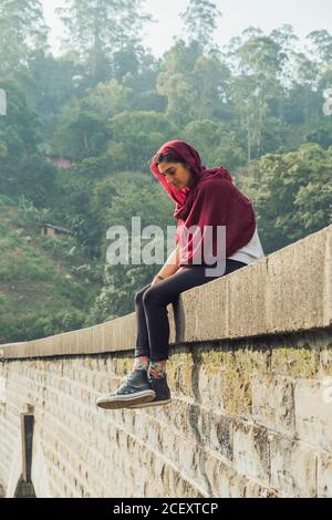 Vue latérale en petit angle sur une jeune femme très attentionnés en tenue active et foulard assis sur le bord en pierre de Vieux pont contre le feuillage tropical vert pendant les vacances au Sri Lanka Banque D'Images