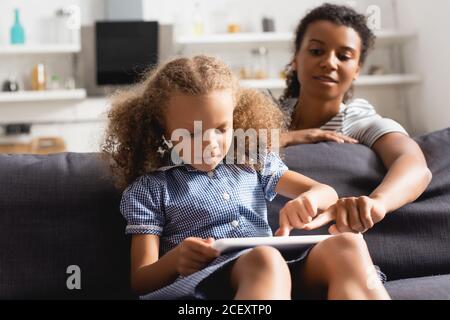 la question sélective de la nounou afro-américaine se met en avant avec le doigt tablette numérique dans les mains d'un enfant assis sur un canapé Banque D'Images