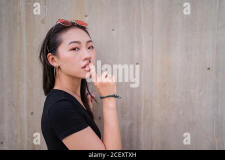 Vue latérale d'une femme asiatique calme debout avec l'index sur les lèvres et en faisant la démonstration de l'affiche en regardant l'appareil photo Banque D'Images