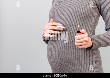 Gros plan d'une cigarette et d'un allume-cigare dans les mains de la femme enceinte sur fond gris. Mauvaise habitude pendant la grossesse. Tabagisme. Banque D'Images