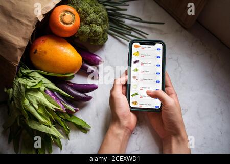 Vue de dessus d'une femme sans visage à la table avec sac en papier avec des produits d'épicerie frais, choisissez des aliments et faites des achats en ligne via l'application sur un smartphone Banque D'Images