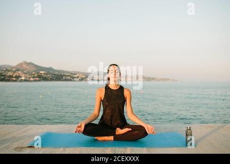 Femme décontractée assise sur un tapis de yoga à Padmasana avec mudra mains et pratique de la pleine conscience avec les yeux fermés sur le fond de paysage marin au coucher du soleil Banque D'Images