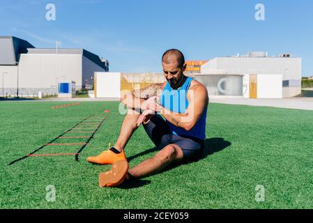 Athlète musclé de sexe masculin assis sur une pelouse verte et vérifiant la forme physique une montre intelligente pour se reposer après un entraînement intensif à l'extérieur Banque D'Images