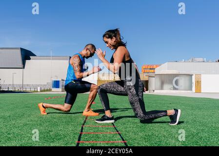 Vue latérale du corps de l'homme et de la femme sportifs qui font des fentes tout en s'exerçant avec l'échelle d'agilité sur une pelouse verte herbeuse sur un terrain de sport Banque D'Images
