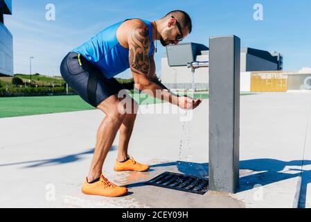 Vue latérale de l'homme sportif musclé tatoué en buvant des vêtements de sport l'eau froide rafraîchissante de la pompe de rue après un entraînement difficile ville Banque D'Images