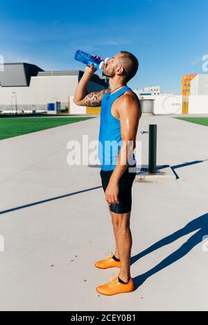 Vue latérale de l'eau potable de l'athlète masculin moueté musclé tatouée de la bouteille en plastique tout en se rafraîchissant après un entraînement intense en milieu urbain rue Banque D'Images