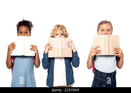 des écolières et des écoliers multiculturels qui couvrent des visages avec des livres et regardent à la caméra isolée sur blanc Banque D'Images