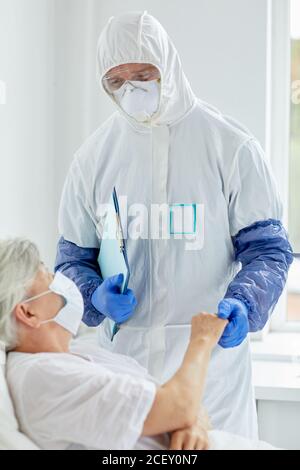 Médecin professionnel portant une combinaison de protection avec des gants pour examiner la santé de femme sénichée au lit dans un service de maladies infectieuses Banque D'Images