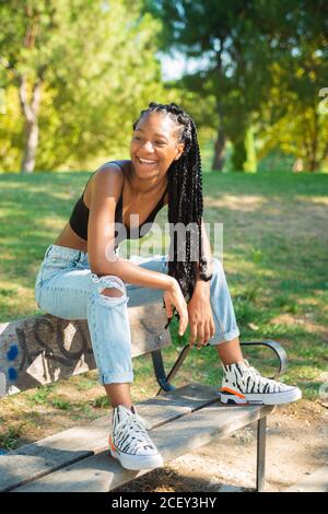 Ensemble de jeunes filles afro-américaines décontractées taille basse tenue tendance assise sur un banc en bois dans un parc verdoyant jour d'été Banque D'Images