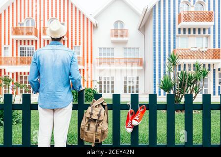 Vue arrière de l'homme debout à la clôture sur Costa Nova avec des baskets et un sac à dos sur la clôture et en regardant les couleurs maisons à rayures Banque D'Images