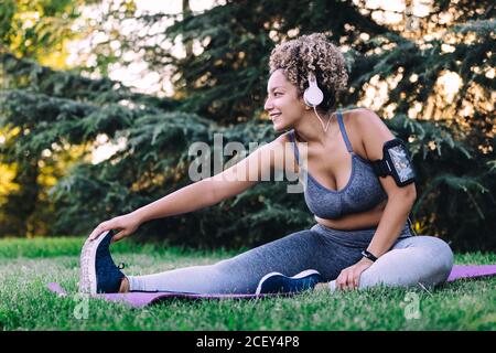 Bonne jeune femme ethnique avec des cheveux bouclés dans l'écoute de sportswear pour écouter de la musique avec un casque ou un smartphone et en étant assis vers l'avant exercice de flexion tout en étirant le corps pendant l'entraînement de fitness en été stationnement
