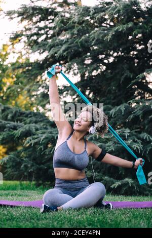 Une femme de taille jeune positive pour tout le corps avec un casque jambes croisées sur l'herbe et bras d'étirement avec bande de résistance pendant l'entraînement physique dans le parc d'été Banque D'Images