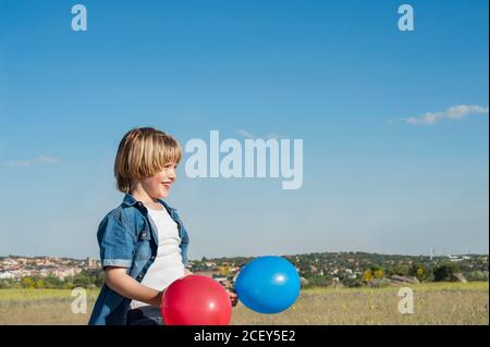 Vue latérale du contenu de l'enfant debout avec ballons d'air colorés dans la prairie et en regardant loin tout en profitant de la journée ensoleillée Banque D'Images