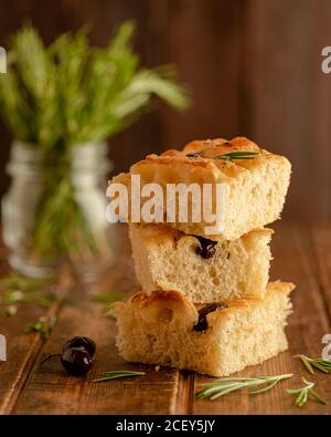 Morceaux de pain de focaccia aromatique fraîchement cuit avec olives noires et romarin frais servi sur un plateau en bois Banque D'Images