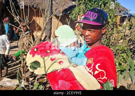 Ilakaka, Madagascar - 30 avril 2019: Inconnu Malgache homme en casquette ajustée tient son bébé sur les mains à côté de la route le jour ensoleillé, simple maison en argile dans l'arrière Banque D'Images