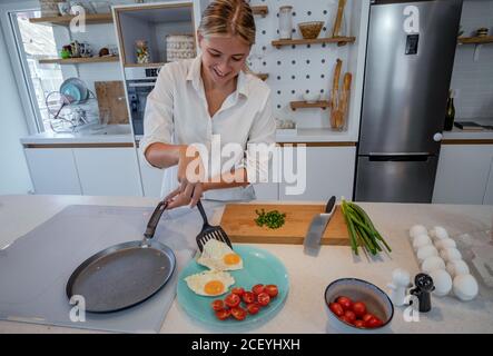 Belle jeune femme cuisine dans une cuisine moderne équipée. Préparation des œufs frits, côté ensoleillé vers le haut. Petit déjeuner à la maison. Banque D'Images