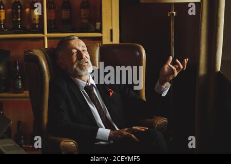 Un vieil homme spectaculaire et agréable regarde avec joie l'appareil photo, s'étire de la paume à la fenêtre, s'assoit confortablement et raconte une histoire joyeuse, posant dans une pièce sombre éclairée, Banque D'Images