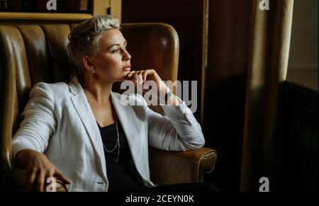 Agréable look ravie femme dans blouson blanc glorieux, avec l'expression réfléchie s'appuie sur l'accoudoir du fauteuil de mode, garde le menton, ne regarde pas Banque D'Images