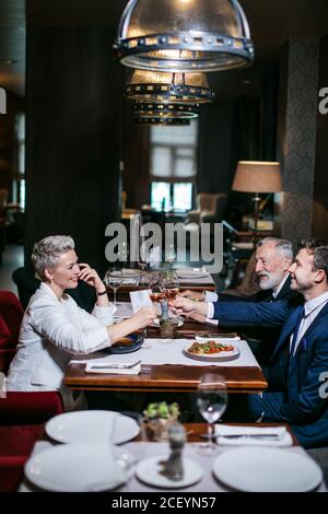 Des partenaires d'affaires joyeux et satisfaits dans des vêtements habillés, asseyez-vous à table, levez des verres à vin avec des visages joyeux, célébrez avec passion l'anniversaire Banque D'Images