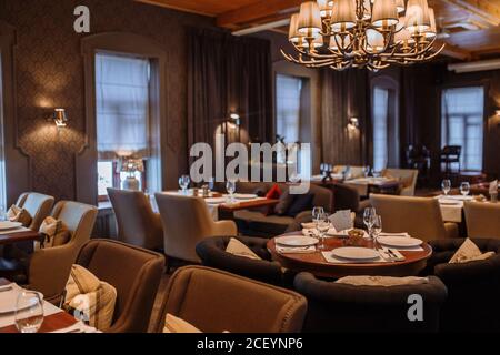 Photo d'un restaurant chic vide avec chaises confortables, lustre vintage au plafond. Des assiettes blanches et des verres à vin se trouvent sur les tables. Ambiance confortable Banque D'Images