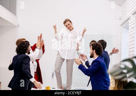 une jeune femme d'affaires caucasienne danse sur table dans un bureau moderne, célébrant l'achèvement réussi de son projet d'affaires, tandis que les collègues soutiennent, a Banque D'Images