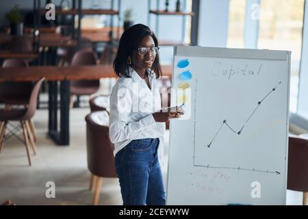 Une femme d'affaires africaine confiante en chemise blanche et en jeans bleus explique comment faire une présentation de tableau de conférence pour les jeunes leaders, partager le bussin Banque D'Images