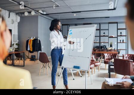 Concept de leadership. Une jeune femme d'affaires en chemise blanche d'afrique se trouve près des diagrammes du tableau de conférence et explique la stratégie d'affaires Banque D'Images