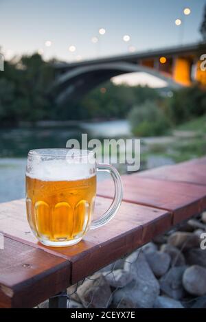 Grand verre de bière pression du Monténégro, illuminé par la lueur du coucher de soleil dans un bar/café au bord de la rivière dans le centre de la capitale du Monténégro. Banque D'Images