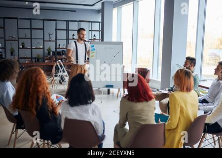 Un jeune entraîneur confiant et enthousiaste donne un discours à ses collègues qui l'écoutent attentivement au bureau, en portant des vêtements décontractés Banque D'Images