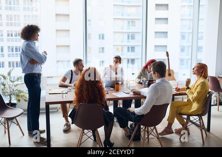 Jeune patron aux cheveux bouclés portant une chemise classique donnant des instructions aux collègues assis sur la table ensemble et l'écouter attentivement, au bureau arrière Banque D'Images