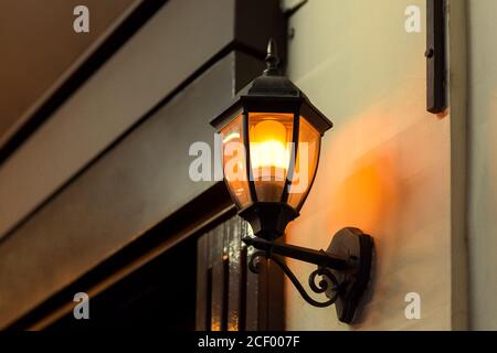 une lanterne en fer noir de style rétro illumine le mur de la façade du bâtiment avec une porte d'entrée en bois, lumière chaude, vue rapprochée du luminaire Banque D'Images