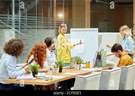 Une jeune femme satisfaite en chemise jaune longue étire la main avec un stylo au tableau noir, montre volontiers le thème de la conférence, fixe à la caméra, calme collègues lo Banque D'Images