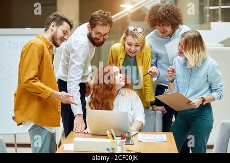 Charmante femme rouge aux cheveux assis à la table avec l'ordinateur sur elle, tourne les mains en arrière, regarde avec sourire le groupe optimiste de designers, demande des conseils, en M. Banque D'Images