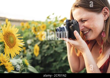 Vue latérale d'une jeune femme souriante photographiant le tournesol à l'aide d'un appareil photo dans la ferme le jour ensoleillé Banque D'Images