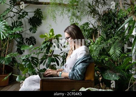 Portrait en gros plan d'une femme souriante gardenier assise sur une chaise en serre, à l'aide d'un ordinateur portable et parlant sur le web entouré de plantes exotiques. Banque D'Images