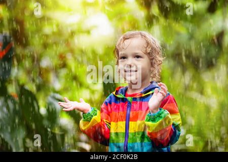 Enfant jouant sous la pluie le jour ensoleillé de l'automne. Enfant sous une forte douche portant une veste imperméable arc-en-ciel. Petit garçon dans le parc des pluies. Activité extérieure de l'automne Banque D'Images