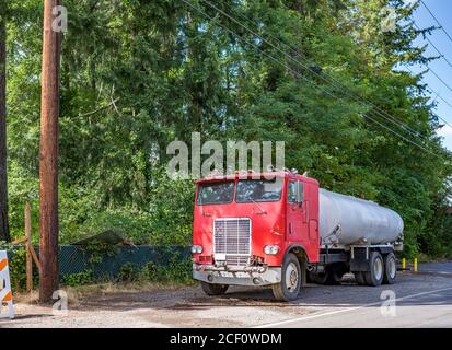 Vieux repeint rouge rétro grand engin de forage semi-camion avec pièces rouillées et remorque-citerne sur la route locale épaule avec arbres verts et clôture Banque D'Images