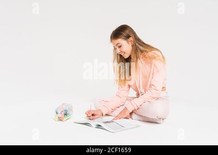Adorable jeune fille souriante avec de longs cheveux dans un pull à capuche rose faire des devoirs isolés sur fond blanc Banque D'Images
