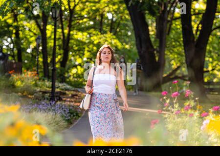 une jeune femme vêtue d'un haut clair et d'une jupe variable marche à travers le parc de la ville, des fleurs défoquées en premier plan Banque D'Images