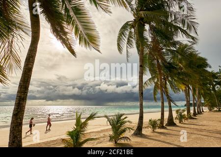 Jeune couple marchant le long de la plage blanche propre avec des cocotiers au coucher du soleil avec ciel nuageux à Boracay Island, province d'Aklan, Visayas, Philippines