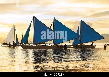 Bateaux à voile bleus attendant les touristes à la rive de la plage blanche sur l'île de Boracay, Philippines avec un coucher de soleil doré dans le ciel et l'eau Banque D'Images