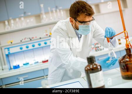 Beau chercheur en vêtements de travail de protection debout dans le laboratoire et flacon d'analyse avec liquide Banque D'Images