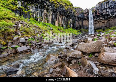 Cascade Svartifoss avec vue sur la rivière en grand angle sur la piste Dans le parc national de Skaftafell en Islande, voir l'eau tomber falaise dans les terres verdoyantes d'été Banque D'Images