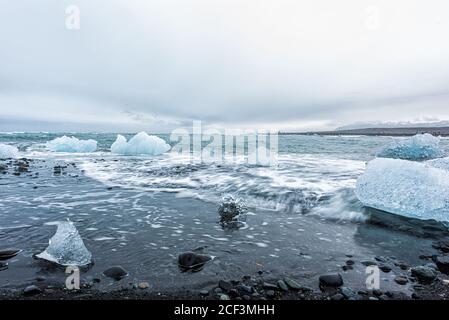 Les vagues sont exposées à une longue exposition sur la glace bleue de l'iceberg flottant Sur des cailloux de sable noir par Jokulsaron lagon lac diamant plage en Islande Banque D'Images